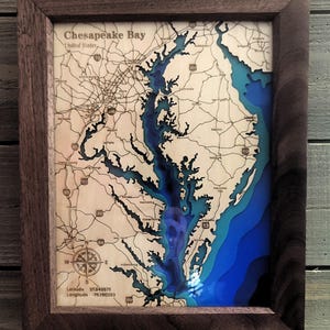 May include: A framed wooden map of the Chesapeake Bay, United States. The map features layered blue and teal water depths, with a detailed depiction of the bay's contours. The frame is a dark brown color, and the map includes text indicating latitude and longitude.