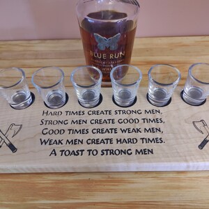 May include: Wooden shot glass holder with a quote about strong men and hard times. The holder has six shot glasses and a space for a bottle. The quote reads: "Hard times create strong men, strong men create good times, good times create weak men, weak men create hard times. A toast to strong men."