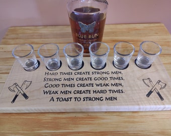 May include: Wooden shot glass holder with a quote about strong men and hard times. The holder has six shot glasses and a space for a bottle. The quote reads: "Hard times create strong men, strong men create good times, good times create weak men, weak men create hard times. A toast to strong men."