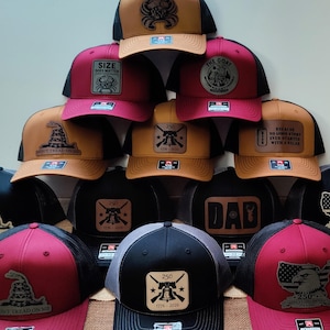 May include: A collection of trucker hats with various designs. The hats feature a range of colors, including black, red, and tan. Some hats display patches with text and graphics, such as "SIZE DOES MATTER," "THE GOAT," and "250 1776-2026."