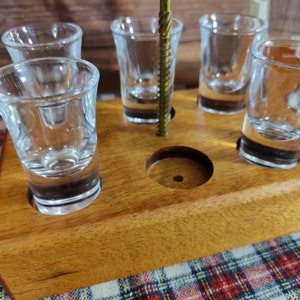 May include: A wooden shot glass holder with five clear shot glasses. The holder is made of light brown wood and has a brass-colored metal accent. The shot glasses are empty and angled slightly.