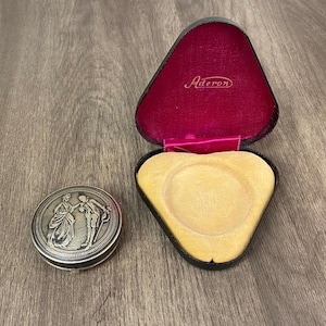 Könnte beinhalten: Ein silberner Kompaktspiegel mit einem detaillierten Relief von zwei Figuren in historischer Kleidung, neben einer schwarzen, dreieckigen Schmuckschatulle mit rosa Innenraum und dem Namen "Aderon" in Gold aufgedruckt.
