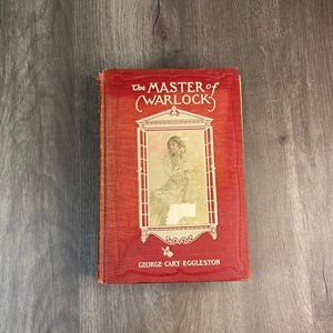 May include: A vintage book titled "The Master of Warlock" with a red cover. The cover features a framed illustration of a woman and the author's name, George Cary Eggleston. The book is placed on a wooden surface.