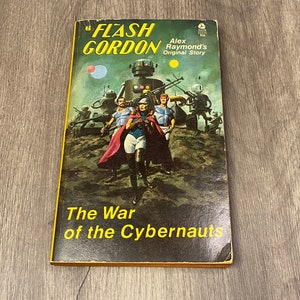 Pode incluir: Um livro de bolso vintage intitulado "Flash Gordon" com o subtítulo "The War of the Cybernauts". A capa apresenta uma cena de ficção científica com personagens em trajes futuristas, robôs e uma capa vermelha. O livro tem uma borda amarela.