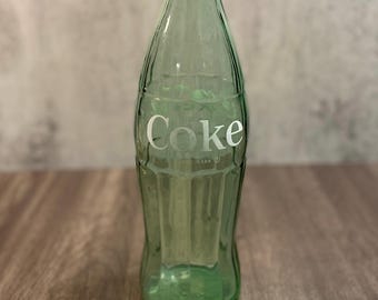 Pode incluir: Uma garrafa de vidro verde-claro de estilo vintage com formato contornado e sulcos verticais. A palavra Coke está gravada em letras brancas na frente, acompanhada pelo texto Trade-mark abaixo. A garrafa possui um gargalo estreito e uma base larga.