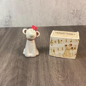 May include: A white ceramic monkey figurine with a red hat and a matching box. The box features musical notes and a cartoon monkey illustration. The text on the box reads "Monkey Shines".