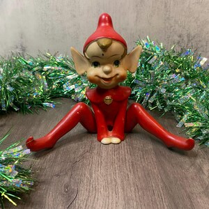 May include: A vintage Christmas elf figurine in a red outfit and hat, sitting with legs spread. The elf has a smiling face, pointed ears, and a gold emblem on its chest. Green tinsel garland surrounds the elf.