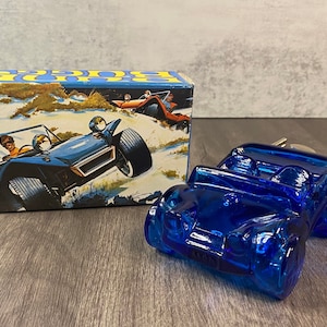 Puede incluir: Una botella de vidrio azul en forma de coche con una caja en el fondo. La caja tiene una imagen de un buggy de dunas azul con dos personas en él.