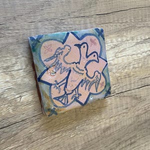 Könnte beinhalten: Eine Keramikfliese mit einem blauen, grünen und rosa floralen Design. Drei stilisierte Vögel sind in Schwarz auf der Fliese gezeichnet, wobei ein Vogel in der Mitte und zwei kleinere Vögel auf jeder Seite sind.