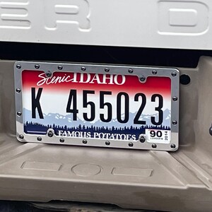 May include: An Idaho license plate with a red, white, and blue gradient background featuring a mountain range and forest silhouette. The plate displays the text Scenic IDAHO, K 455023, and FAMOUS POTATOES, enclosed in a silver metal frame with rivets.