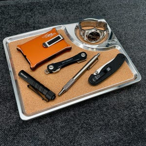 May include: A rectangular silver metal valet tray with a cork inlay containing an orange wallet with a carbon fiber clip, a silver wristwatch, a black KeySmart key organizer, a silver metal pen, a black Olight flashlight, and a black pocket knife.