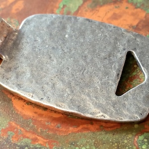 May include: A silver pendant with a triangular cutout and a loop for attaching a chain. The pendant has a textured surface and a slightly rounded shape.