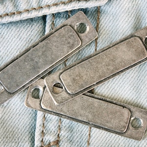 Puede incluir: Tres etiquetas de metal plateado con bordes redondeados y agujeros para cuerda o cinta. Las etiquetas están sobre un fondo de mezclilla azul claro.