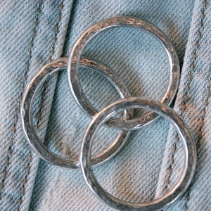 May include: Three silver rings, each with a hammered texture, are stacked on top of each other. The rings are on a blue denim background.
