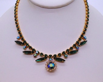 May include: A gold-toned necklace with emerald green and iridescent blue-green rhinestones. The necklace features a central floral pendant and a delicate chain, displayed on a white jewelry bust.