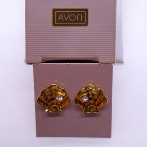 May include: A pair of gold-tone shell-shaped earrings featuring a single clear rhinestone accent on each piece. The earrings are attached to a mauve-colored display card with the brand name AVON printed in a rectangular box at the top.
