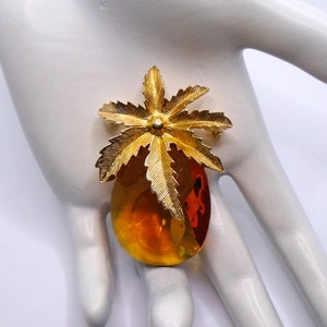 May include: A gold tone brooch featuring a stylized palm tree with a large amber colored glass stone.
