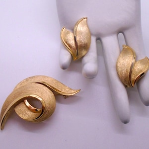 Peut inclure: Un ensemble de bijoux dorés composé d'une broche et d'une paire de boucles d'oreilles. La broche est un design abstrait en spirale, tandis que les boucles d'oreilles sont de simples formes de feuilles.