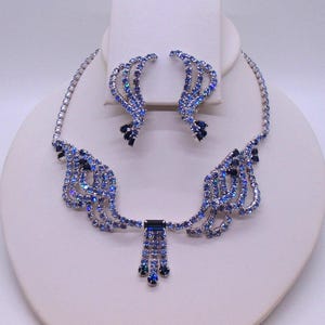 May include: A blue rhinestone jewelry set, including a necklace and matching earrings. The necklace features a central pendant with dangling rhinestones and a delicate chain. The earrings have a curved design. The set is displayed on a white jewelry stand.