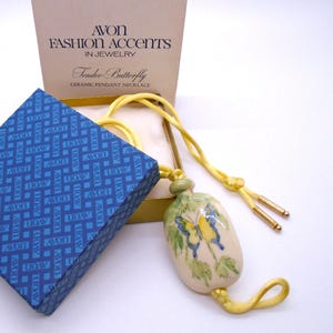 May include: A ceramic pendant necklace featuring a yellow and blue butterfly design on a cream-coloured oval pendant. The necklace includes a yellow cord with gold-tone metal tips, presented in a blue patterned box and a cream-coloured Avon box.