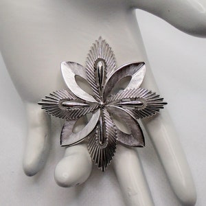 May include: A silver-toned brooch in the shape of a stylised flower with six points. The flower has a textured, slightly raised design.