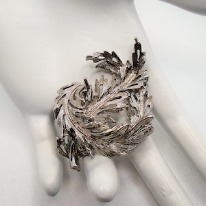 Puede incluir: Un broche plateado en forma de hoja de helecho estilizada.