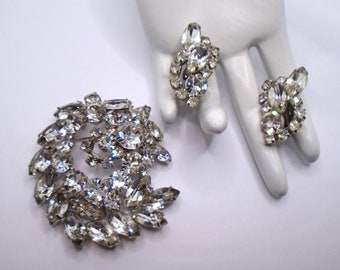 Può includere: Un set di gioielli vintage in strass con una spilla e un paio di orecchini. I gioielli sono realizzati in strass trasparenti incastonati in metallo argentato. La spilla ha un design a spirale e gli orecchini hanno un design a grappolo.