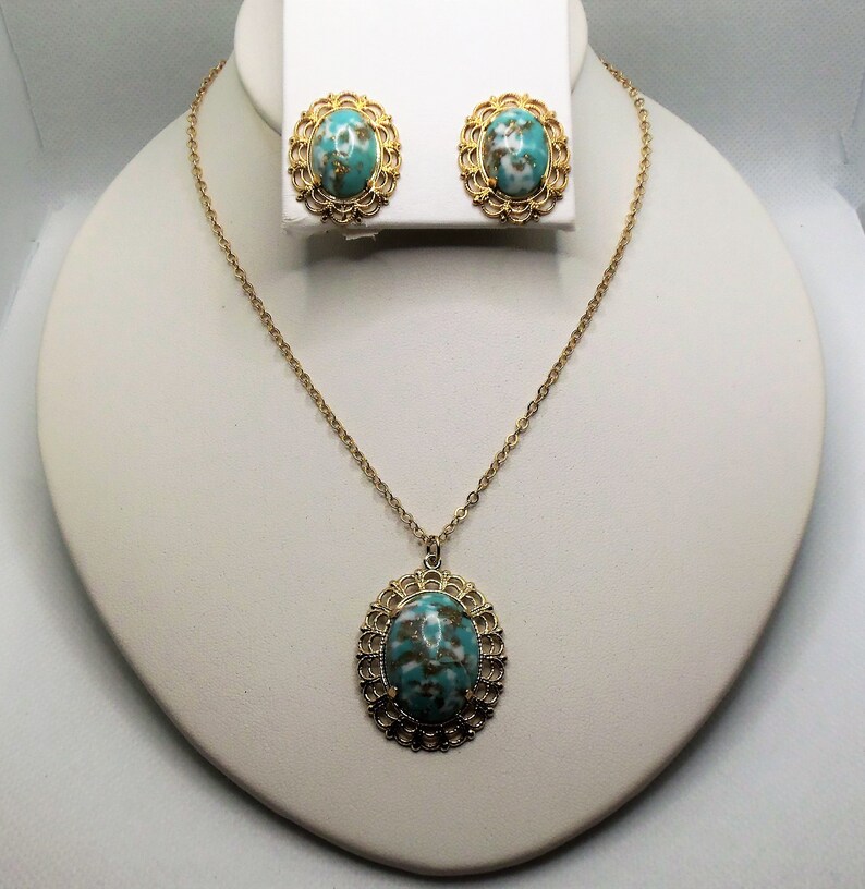May include: A gold necklace and earrings set with a turquoise and gold oval pendant and matching earrings. The pendant and earrings have a delicate filigree design.