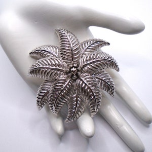 Puede incluir: Un broche plateado en forma de palmera estilizada con hojas detalladas. El broche tiene un pequeño centro redondo plateado.