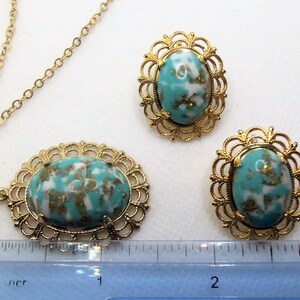 May include: A gold chain necklace with a turquoise and gold oval pendant. The pendant has a delicate filigree design. Matching earrings with the same design are also included.