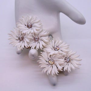 May include: White daisy-shaped earrings with rhinestone centers. The earrings are made of a textured material and are arranged on a white hand-shaped display. The earrings have a vintage aesthetic.