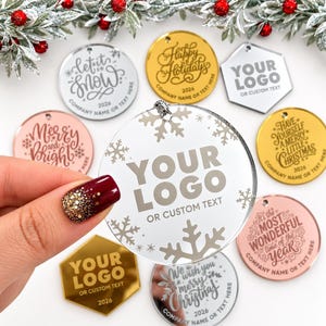 May include: A collection of circular and hexagonal acrylic ornaments in gold, silver, rose gold, and clear finishes. Each piece features engraved holiday text or a placeholder for a custom logo and text, including phrases like Let it Snow.