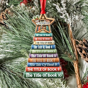 May include: A Christmas tree-shaped ornament featuring a stack of ten books with the text The Title Of Book 1 through 10, topped with a star containing the name Sophia and a bookshelf graphic, suspended by a red ribbon for hanging on a tree.