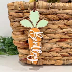May include: A decorative carrot-shaped tag with the name "Bentley" in white script. The tag features an orange carrot body, green leaves, and a brown string. It is hanging on a woven basket.