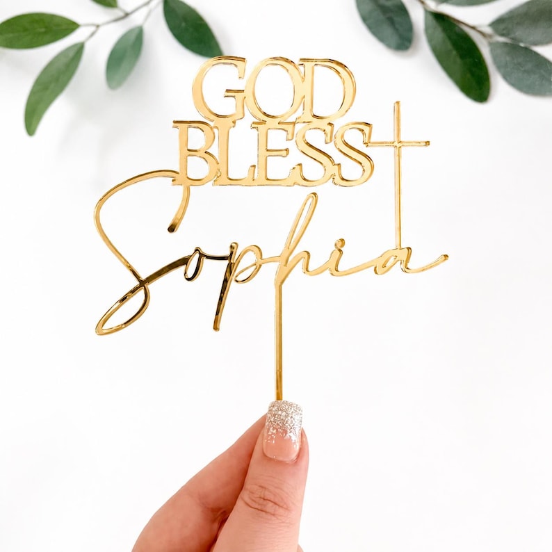 May include: A gold-colored acrylic cake topper featuring the words "GOD BLESS" in a bold, uppercase font above the name "Sophia" in a cursive script. A vertical cross symbol is integrated into the right side of the design, extending from the text.
