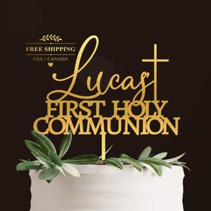 Pode incluir: Um topo de bolo dourado com o nome Lucas em letra cursiva, seguido pelo texto FIRST HOLY COMMUNION em uma fonte negrito sem serifa, com uma cruz integrada ao design, posicionado sobre um bolo branco com folhas verdes.