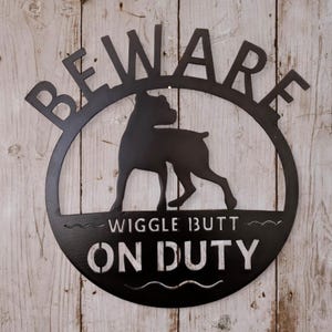 May include: A circular black metal wall sign featuring a silhouette of a dog in the center. The text BEWARE is arched across the top, while the words WIGGLE BUTT ON DUTY are printed in a stencil-style font across the bottom half of the sign.