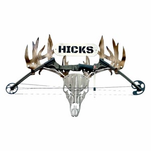 May include: A camouflage-patterned deer skull mount with a bow and arrow attached. The bow is black and green, and the arrow is silver. The skull mount has a wooden plaque above it with the name "HICKS" written in black letters.