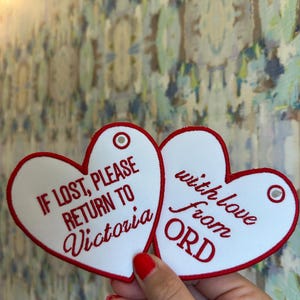 May include: Two white heart-shaped tags with red stitched borders and metal eyelets. The left tag reads, IF LOST, PLEASE RETURN TO Victoria, and the right tag reads, with love from ORD, both in red lettering.