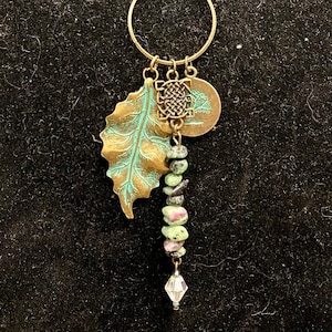 May include: A gold chain necklace with a circle pendant, a leaf charm, a knotted charm, and a round charm with a tree design. The necklace also features a strand of green and pink gemstone beads with a crystal pendant.