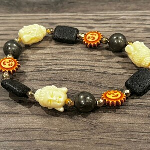 May include: A beaded bracelet featuring cream-colored Buddha head beads, black lava rock beads, and orange sun-shaped beads. The bracelet also includes dark gray and gold-colored beads, strung together on a stretchy cord.