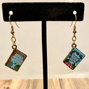 Puede incluir: Un par de pendientes de oro con encantos de libros en miniatura. Los encantos presentan las portadas de "Alice's Adventures in Wonderland" y "Through the Looking Glass".