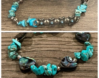 May include: Two turquoise stone bracelets. One features silver-toned flower accents and rose-shaped beads. The other has heart-shaped abalone shell beads. Both bracelets are on a wooden surface.