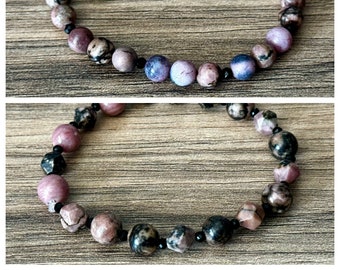 May include: A beaded bracelet featuring a mix of round and faceted stones in shades of dusty pink, mauve, dark gray, and black. The design alternates larger, textured stone beads with small, smooth, black spacer beads throughout the circular strand.