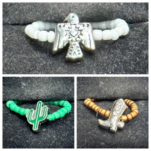 Può includere: Tre anelli di perline con ciondoli diversi. Il primo anello ha un ciondolo di aquila d'argento, il secondo anello ha un ciondolo di cactus verde e il terzo anello ha un ciondolo di stivale d'argento.