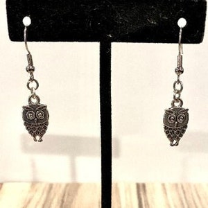 May include: A pair of silver owl dangle earrings on a black display stand. The owls have detailed features and are hanging from silver chains.