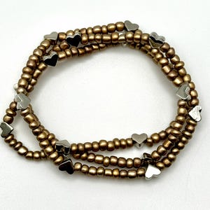 May include: A gold beaded bracelet with silver heart charms. The bracelet is layered and has a delicate look.