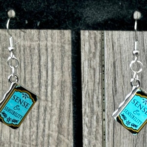 May include: A pair of silver earrings with a blue and black book charm. The book charm features the text "Sense & Sensibility".