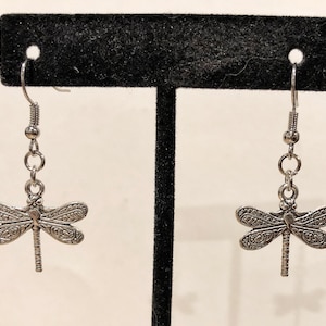 May include: A pair of silver dragonfly earrings with intricate details. The earrings are hanging from a silver hook.