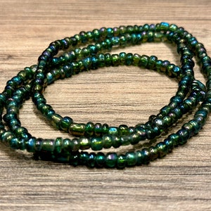 May include: Strands of iridescent green seed beads, arranged in a circular pattern. The beads have a dark green base color with hints of blue and gold, creating a shimmering effect. The beads are small and uniform in size.